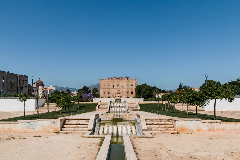 Historic building in the background with trees and landscaped pathways. Steps and water features in the foreground under a clear sky.