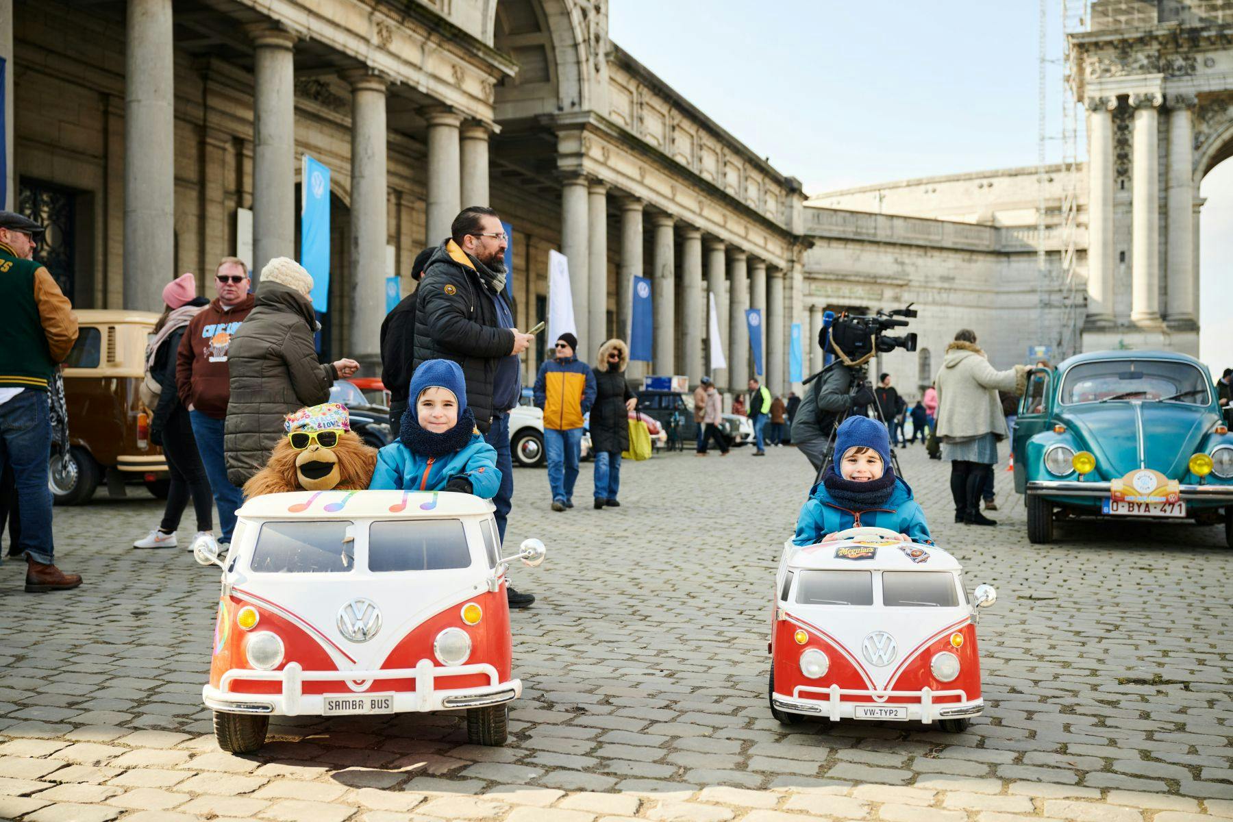 Enfants conduisant des mini-bus VW dans une rue pavée, avec des adultes et des colonnes de bâtiments historiques à l'arrière-plan.
