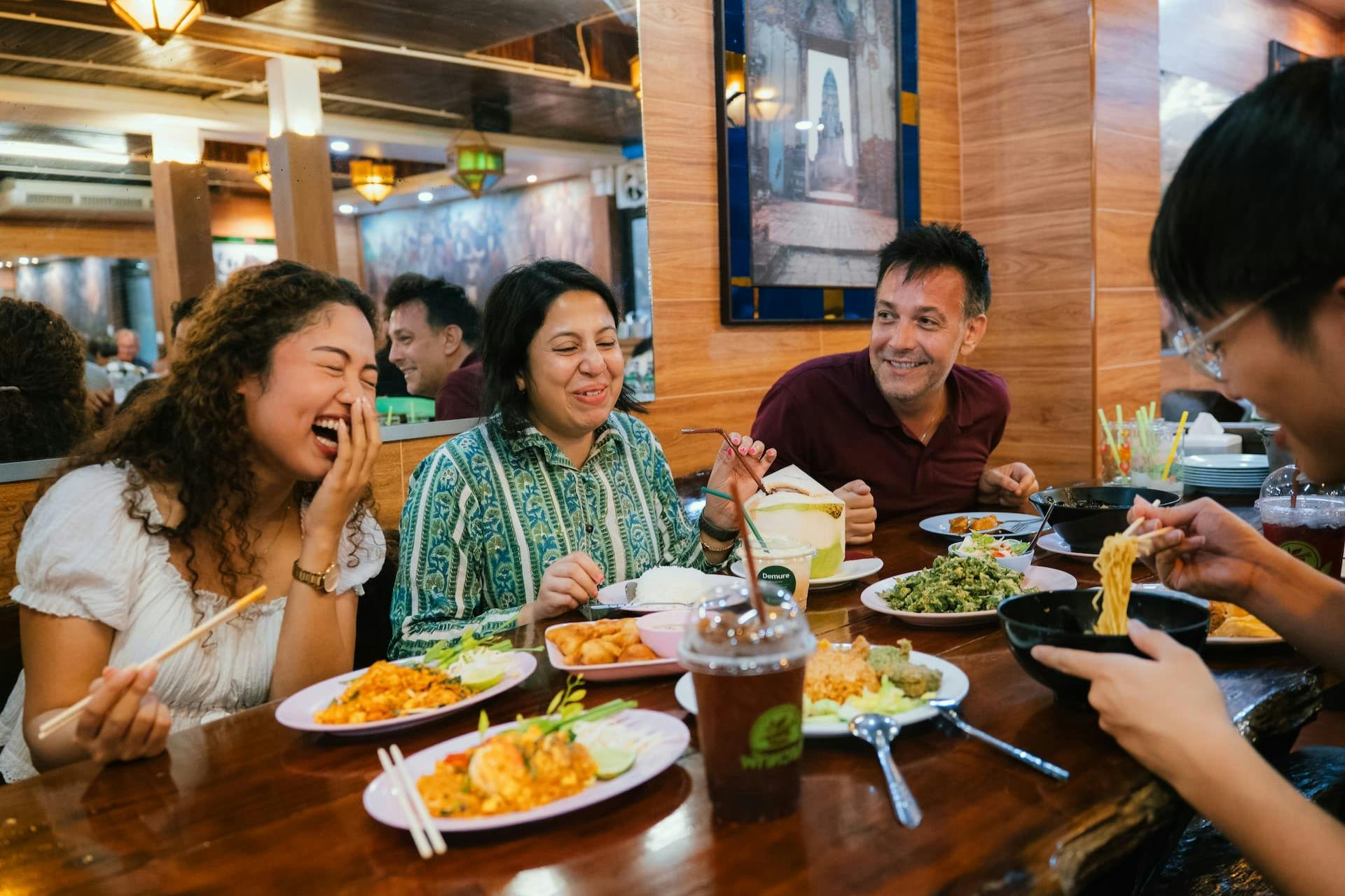 Tre personer sidder ved et bord på en restaurant og smiler og nyder et måltid med forskellige retter foran sig.