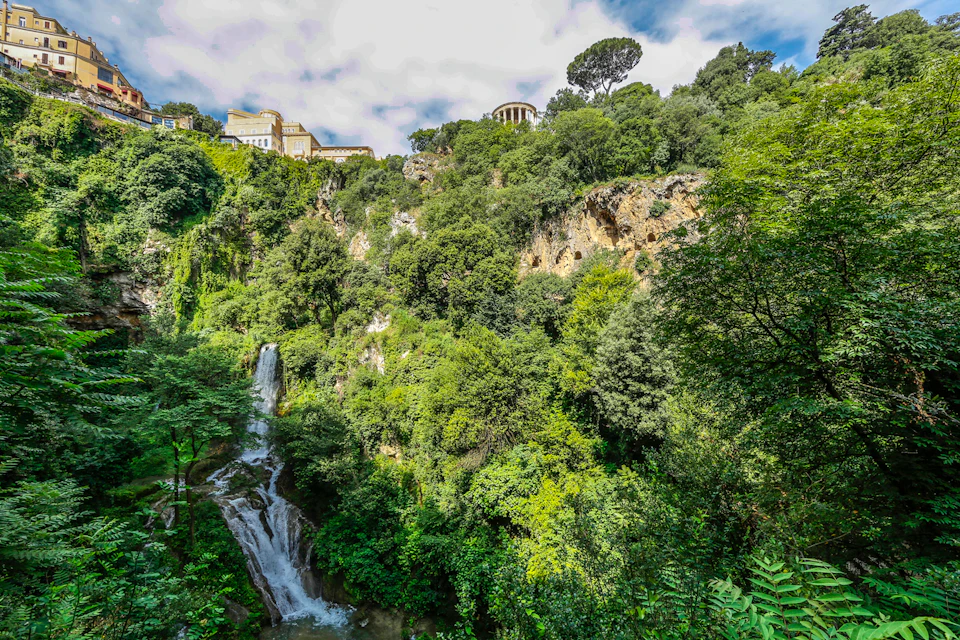 A waterfall cascades down lush, green hills with buildings perched at the top under a partly cloudy sky.