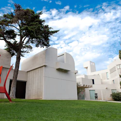 En modern vit byggnad med rundade kanter, en röd abstrakt skulptur, träd och en gräsbevuxen gräsmatta under en blå himmel med moln.