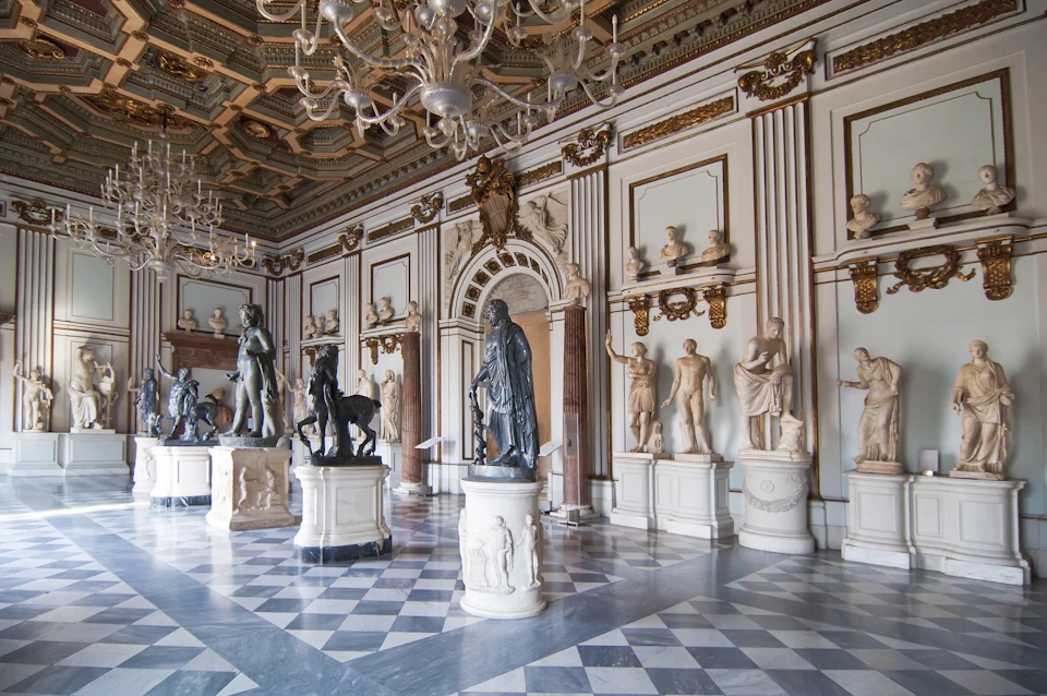A grand hall with marble statues, ornate chandeliers, decorative columns, and a checkered floor.