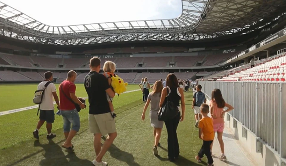 People walking on a large, empty football field inside a stadium. A man carries a child holding a yellow stuffed toy.
