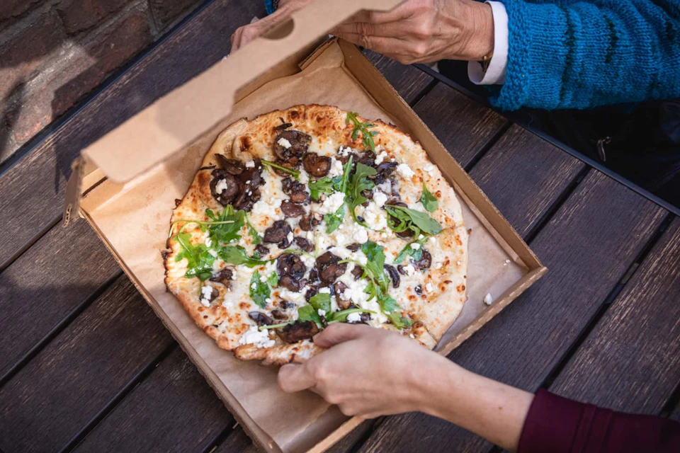 Two hands opening a box of pizza topped with mushrooms, arugula, and cheese. The box sits on a wooden table.