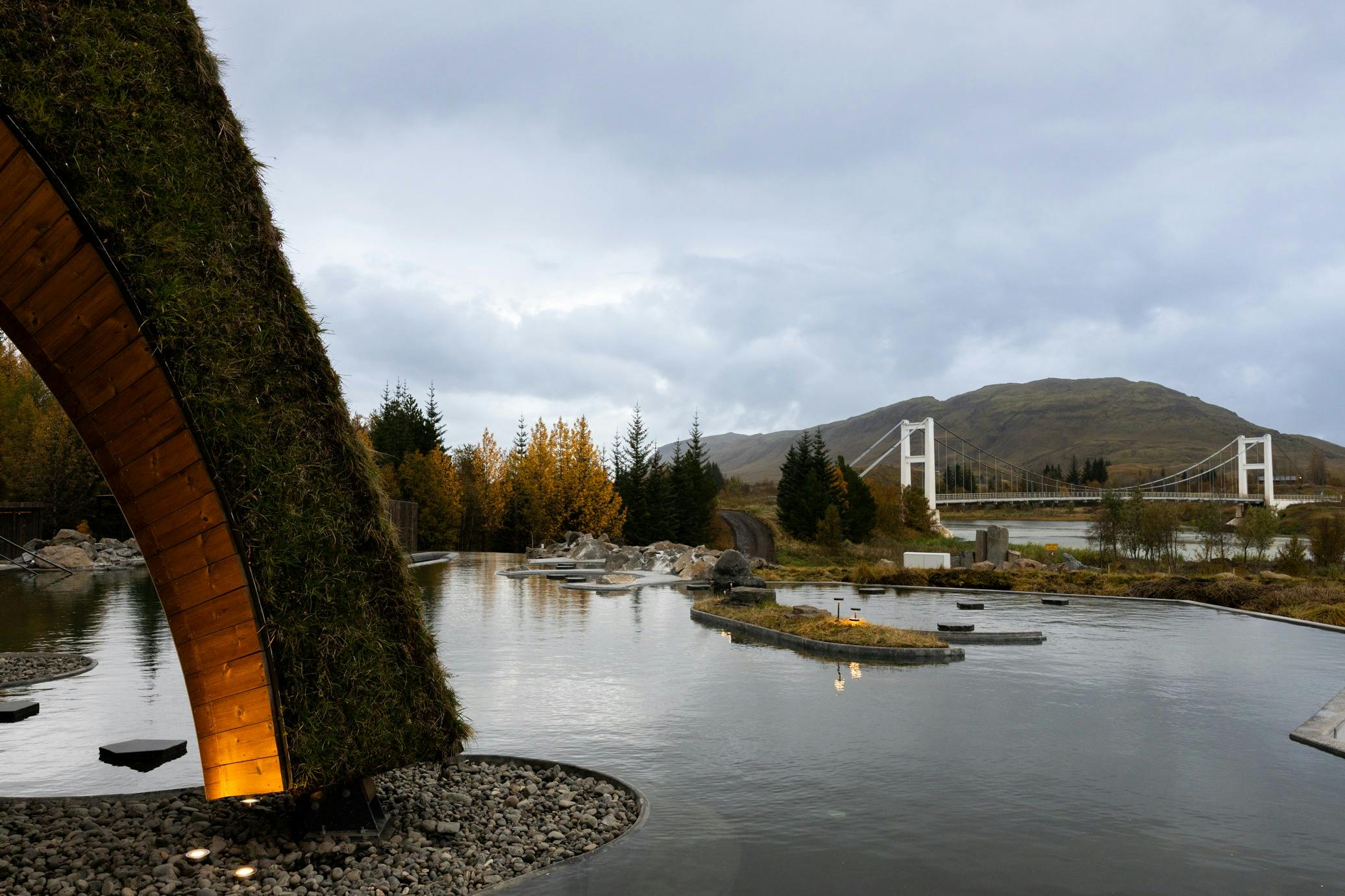 Grass-roofed structure by a calm body of water with small islands, trees, a bridge, and mountains under a cloudy sky.