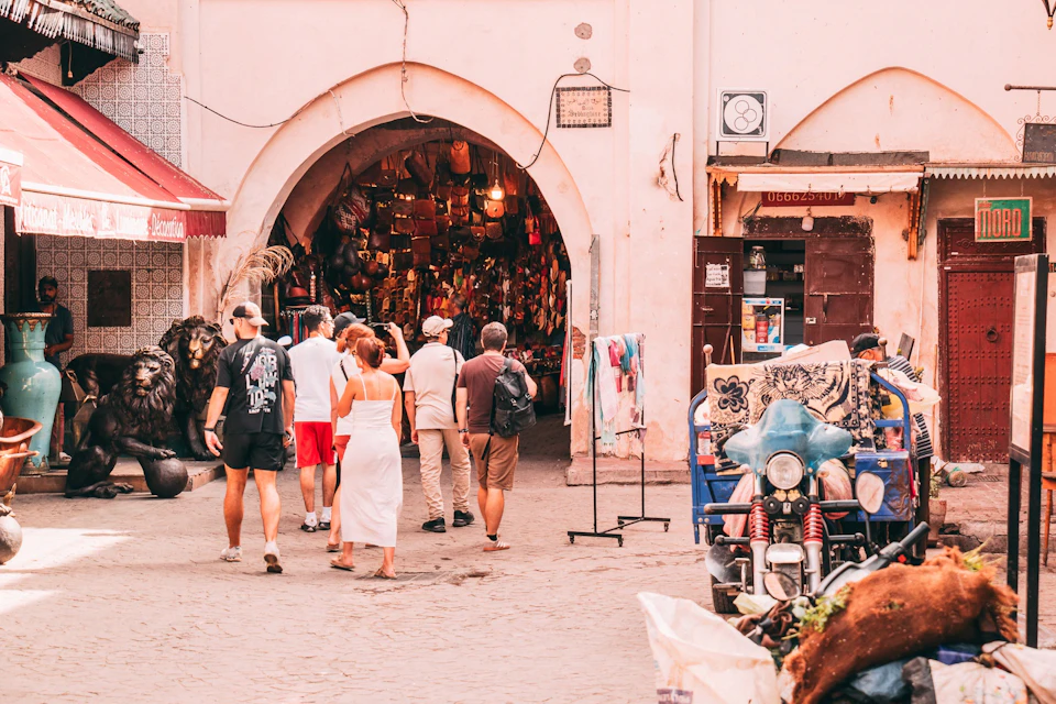 A group of tourists walks toward an open-air market stall filled with colorful textiles, baskets, and souvenirs.