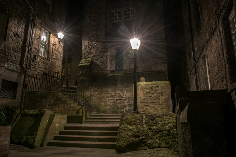 A dimly lit stone alleyway at night with stairs, streetlights, and historic brick buildings.