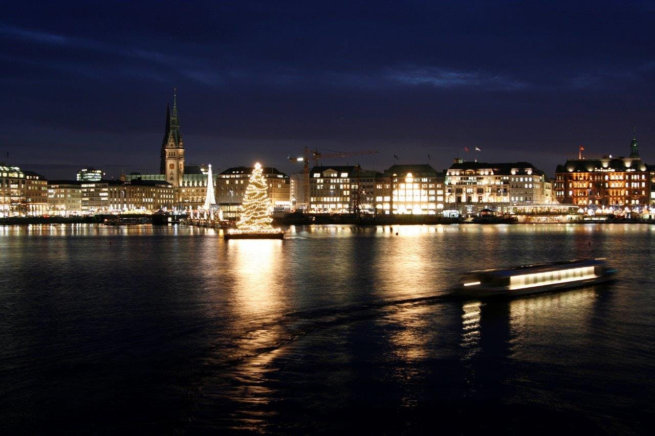 Un paesaggio urbano di notte con edifici illuminati, un'importante torre dell'orologio e un grande albero di Natale che si riflette in un fiume.