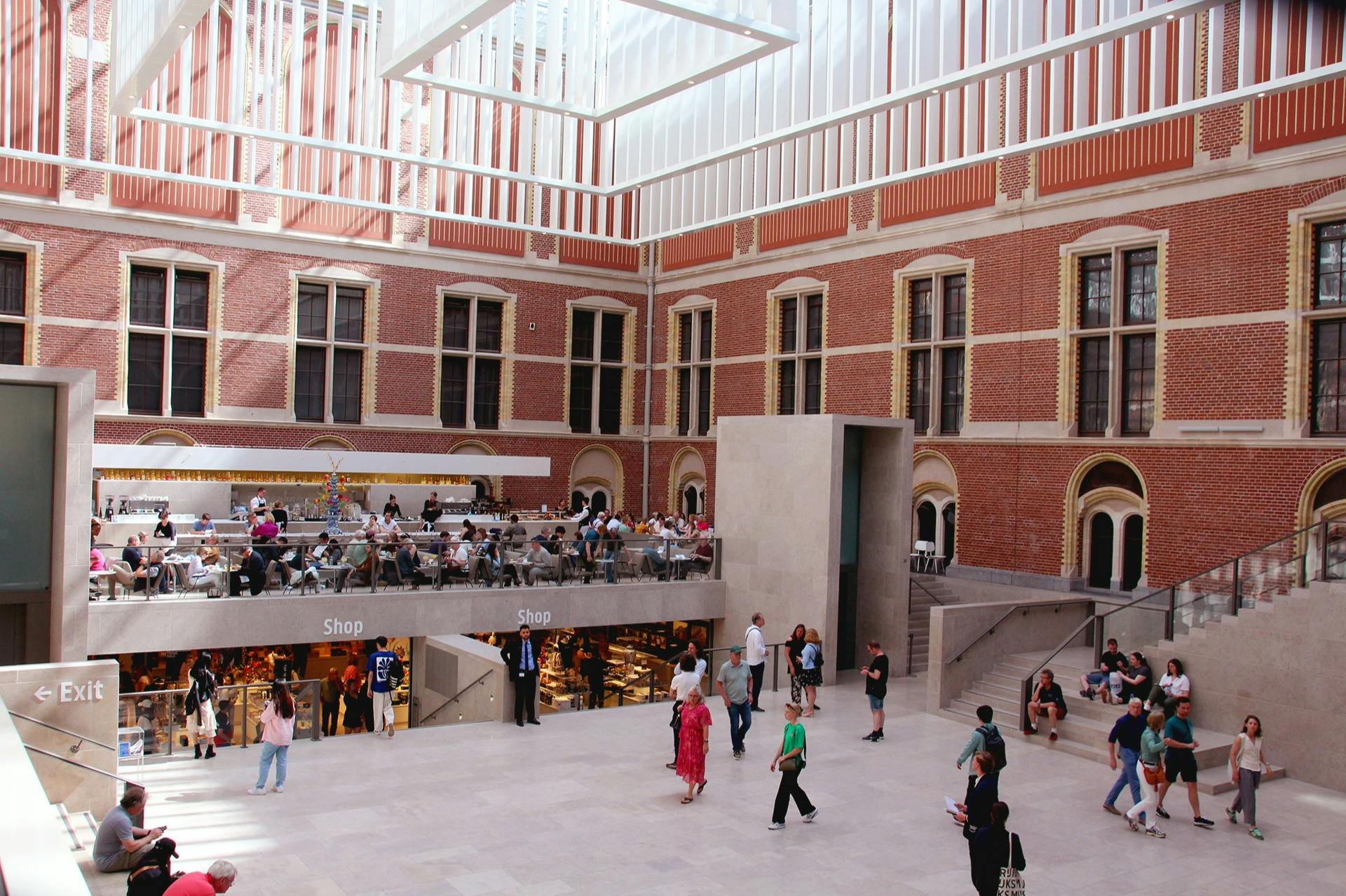 A spacious, modern atrium with a mix of red brick and glass elements, people walking, and a café and shops on the upper level.