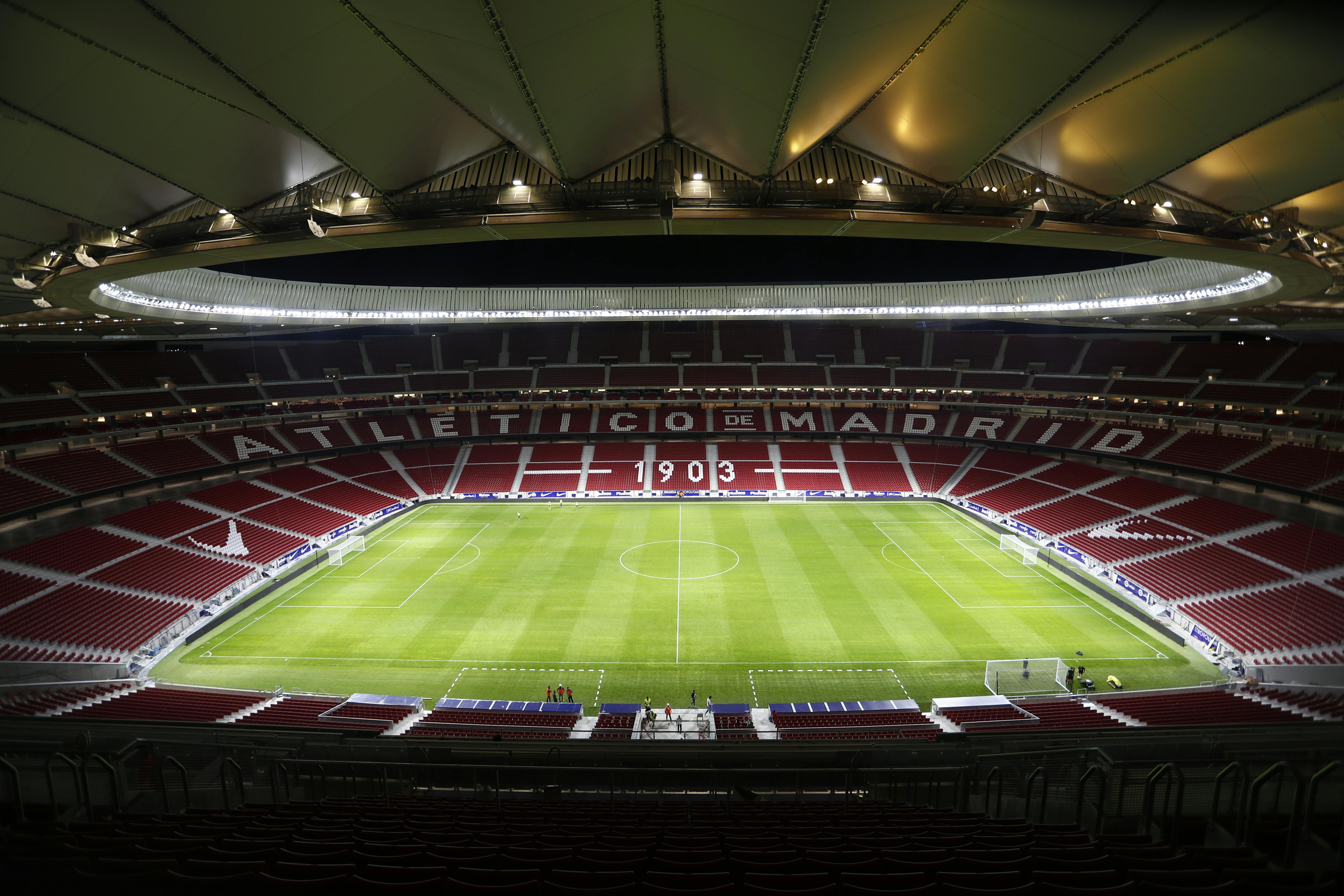 Empty football stadium with red seats, green pitch, and "Atletico de Madrid" written on the stands, under a roof with lights.