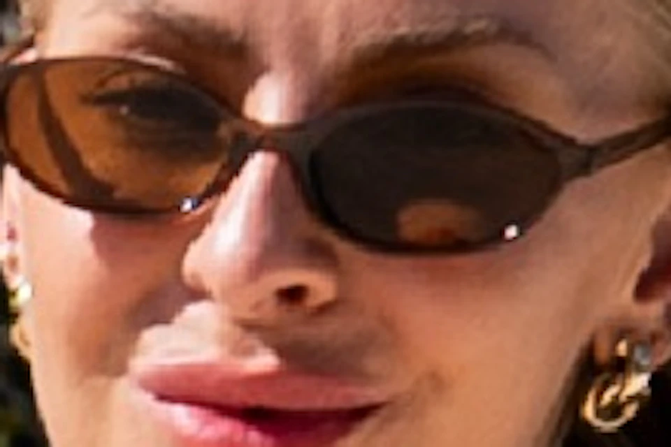 A woman rides a red bicycle on a sunny, paved path with trees in the background. She wears sunglasses and casual summer clothing.