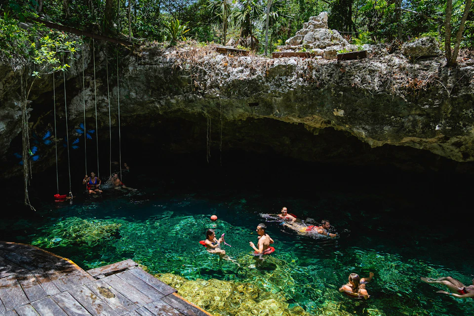 People swimming and relaxing in a clear, blue natural pool surrounded by rocky terrain, vegetation, and an overhead cliff.
