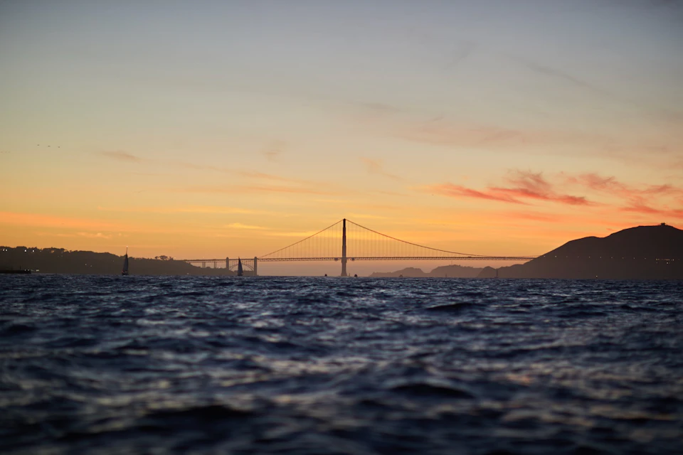 A view of the Golden Gate Bridge at sunset, with rippling water in the foreground and an orange-yellow sky behind.
