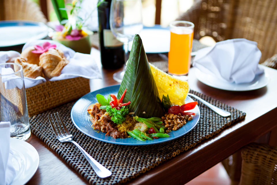 Plate of Indonesian food with a banana leaf cone, vegetables, sambal, crackers, and orange juice on a woven placemat.