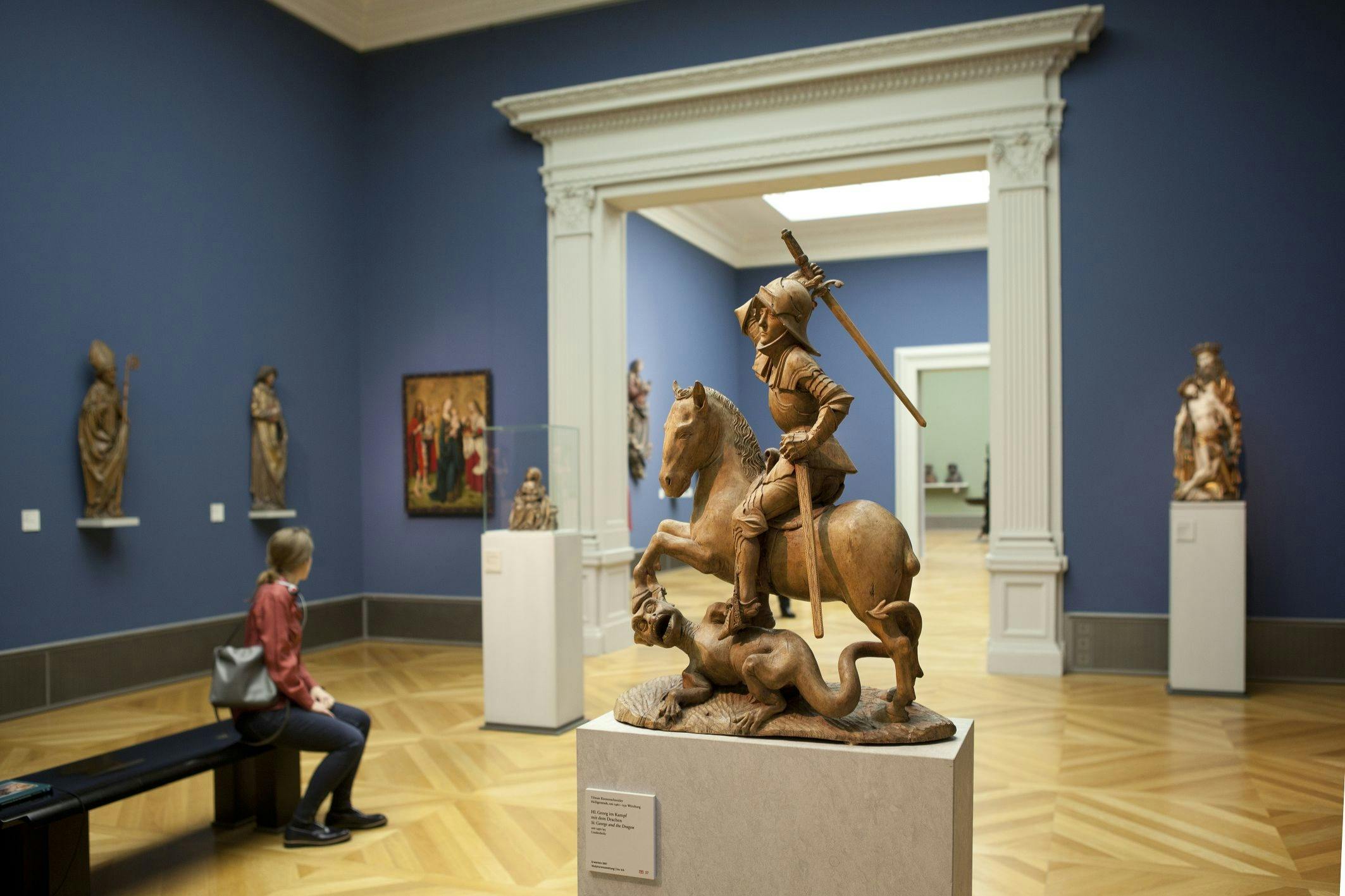 Una sala de museo con una escultura de un caballero a caballo, un visitante sentado y obras de arte expuestas en paredes azules.
