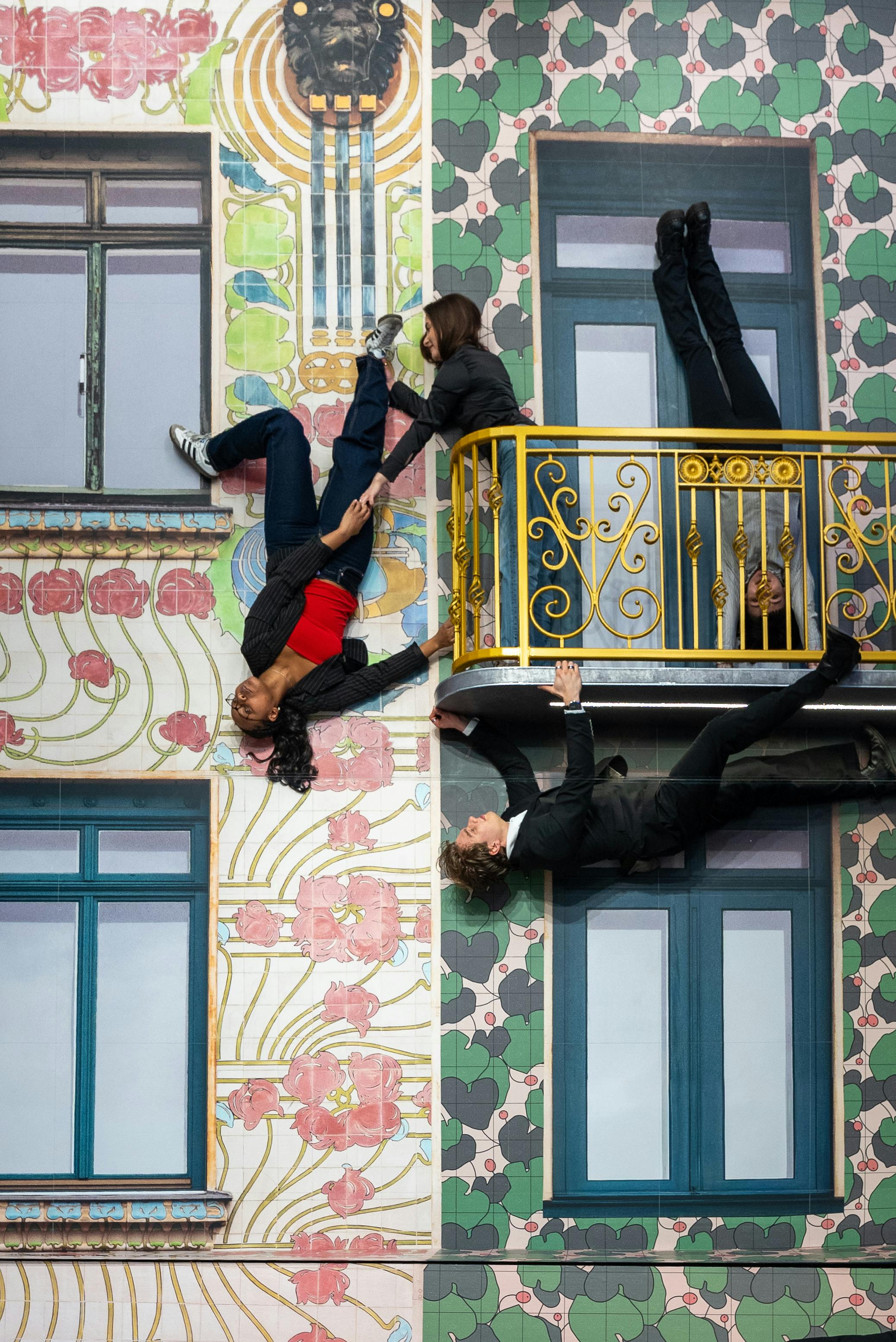 Folk synes at trodse tyngdekraften på en farverig bygningsfacade med vinduer, en balkon og blomstermønstre.