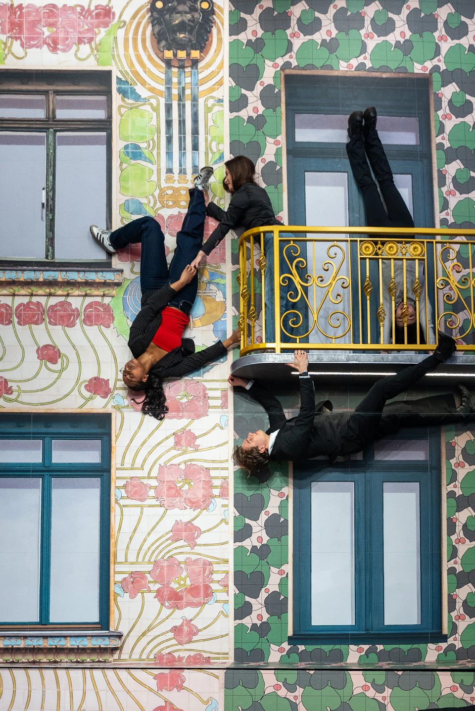 People appear to defy gravity on a colourful building facade with windows, a balcony, and floral patterns.