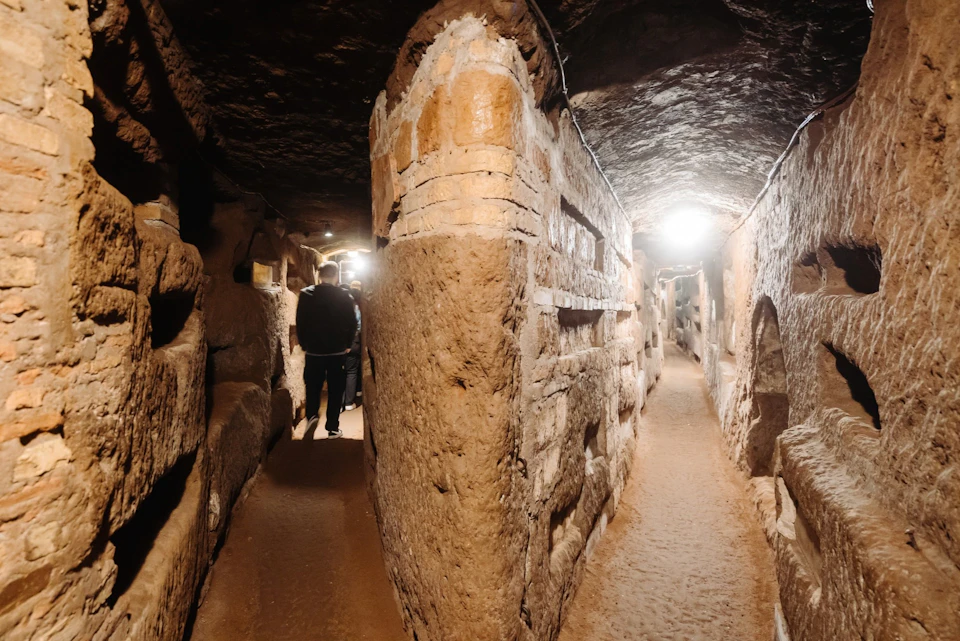 A dimly lit underground tunnel with stone walls and arched passages, featuring a person walking in the distance.
