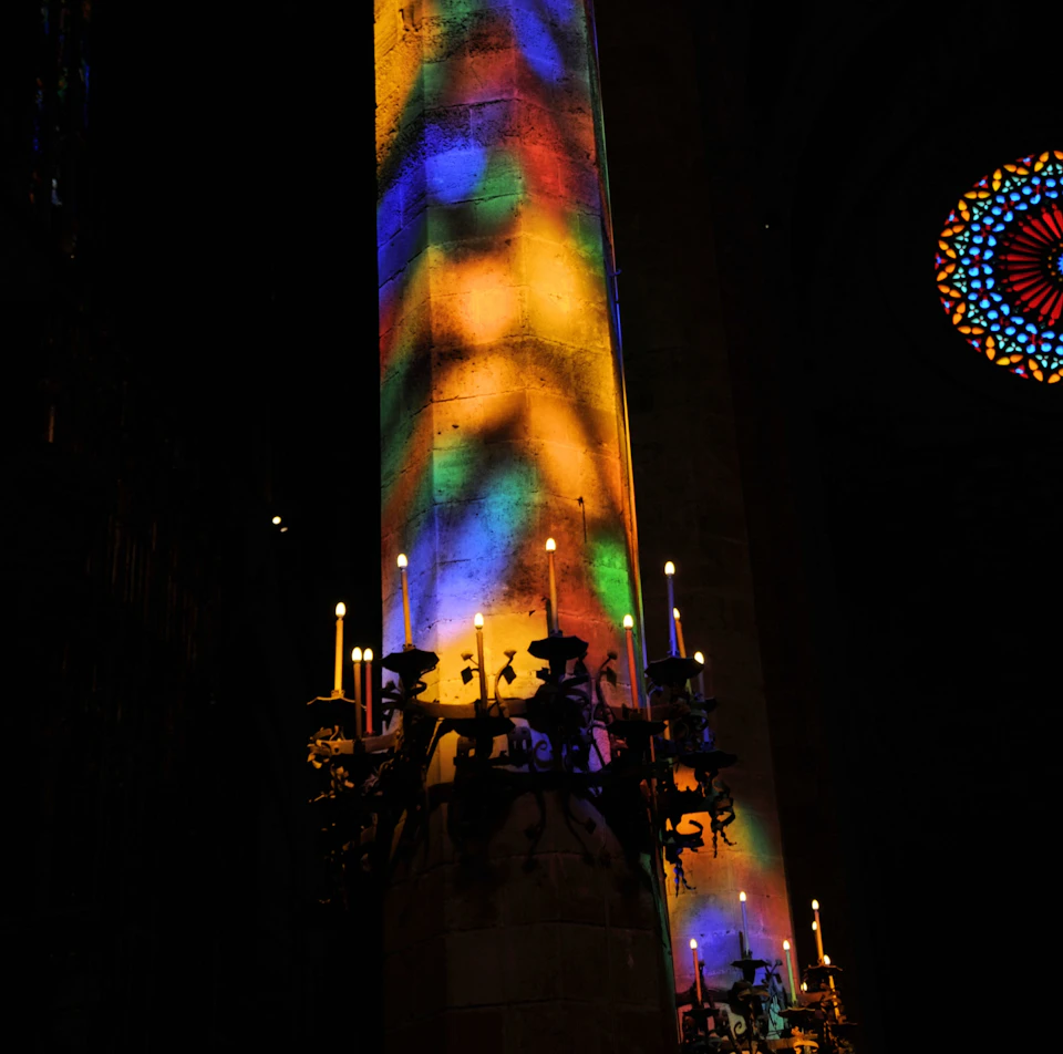 Colorful light patterns illuminate a large pillar in a dimly lit church, with candles on ornate candle holders.
