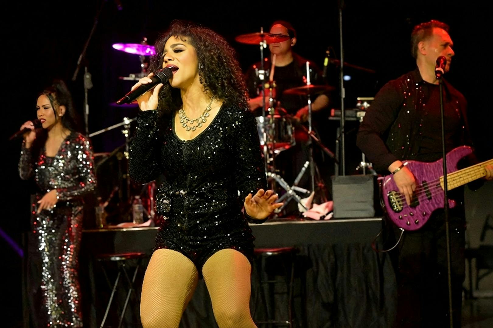 A female singer in a black sequined outfit performs on stage with a live band, including a backup singer and a drummer.