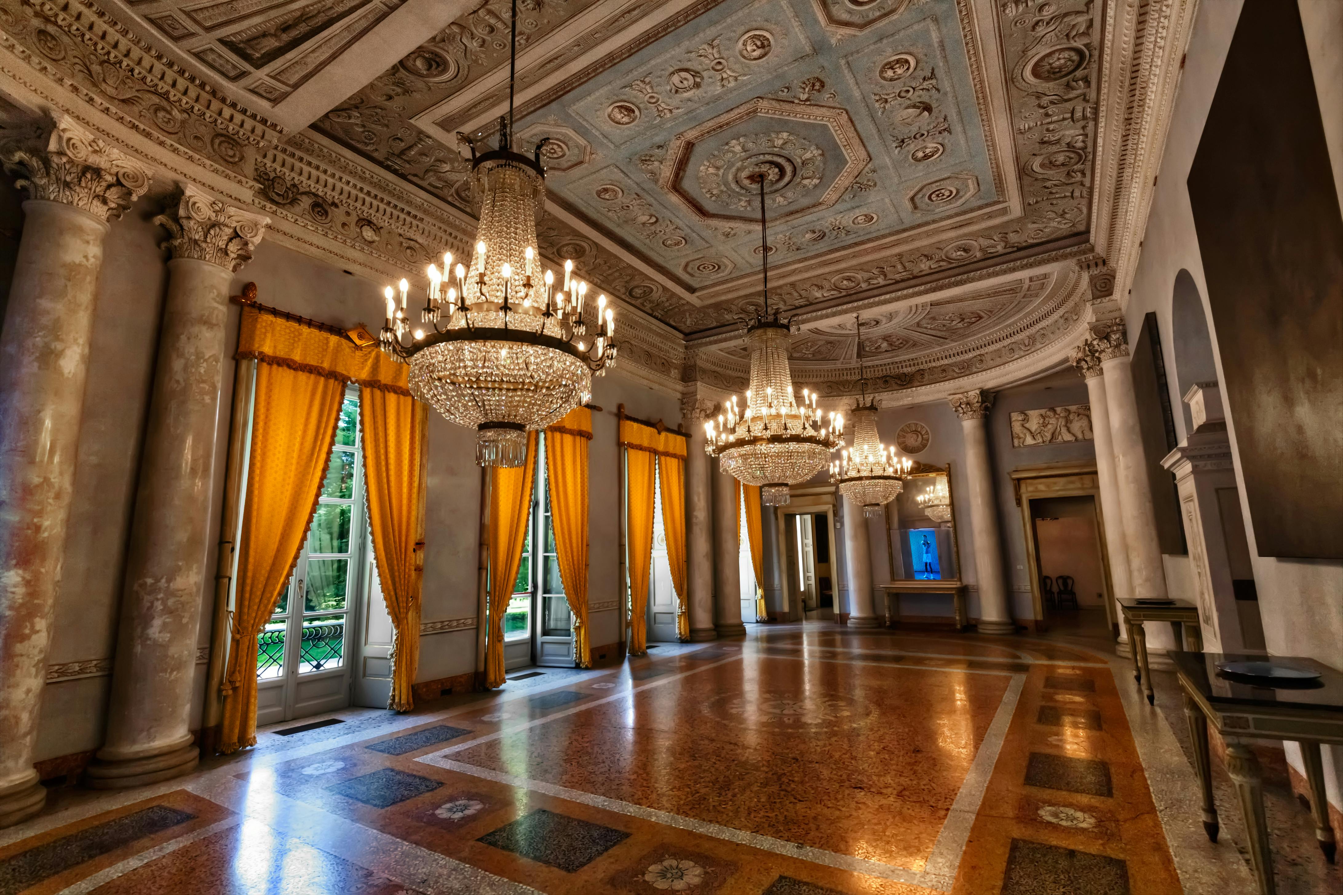 Une salle de bal ornée de motifs complexes au plafond, de grands lustres et de hautes tentures jaunes couvrant les grandes fenêtres.