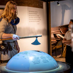 En person i historisk kostym håller en yxa och interagerar med en grupp nära en världsutställning i en museumsmiljö.