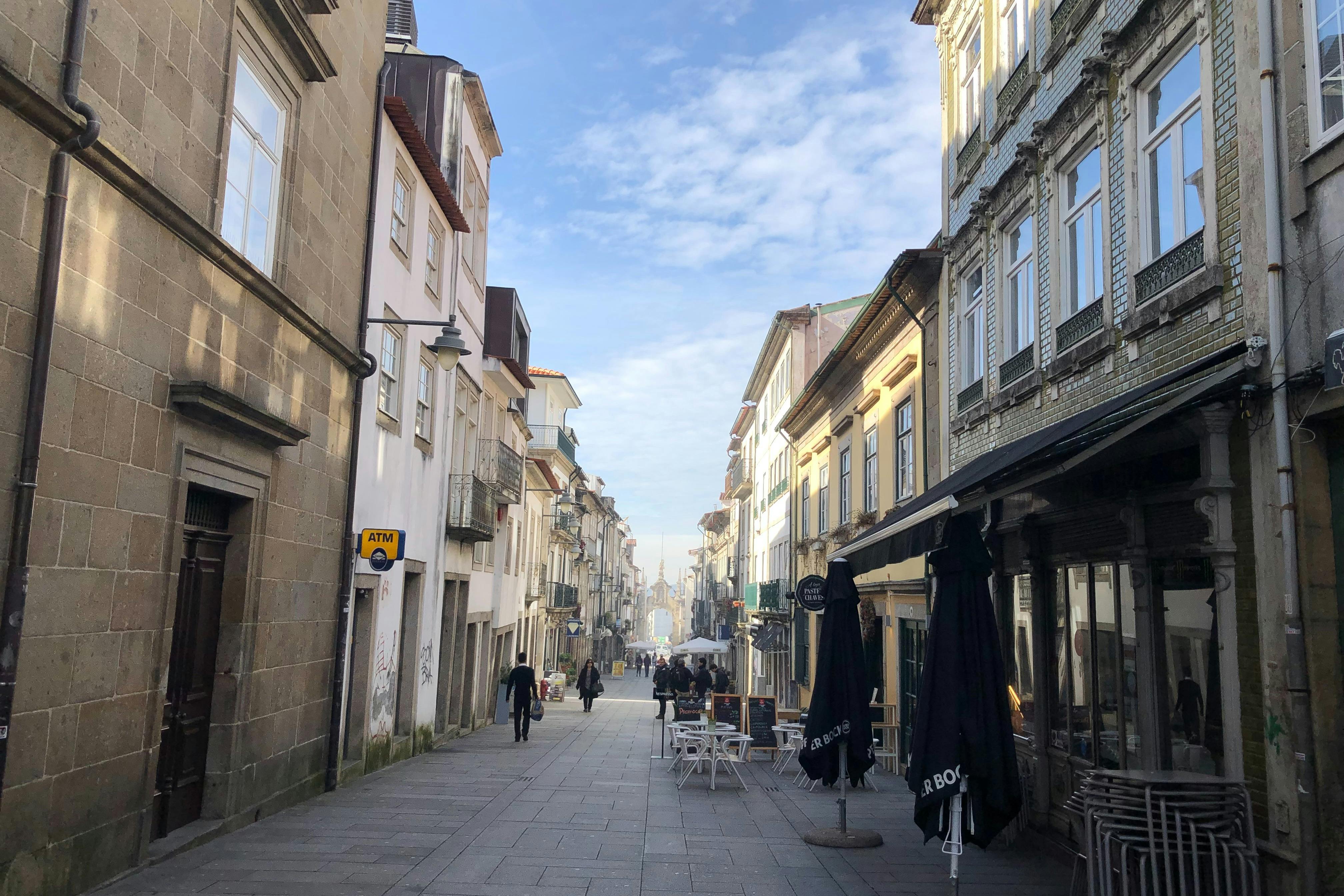 A narrow cobblestone street lined with old buildings, outdoor cafe tables with umbrellas, and a few people walking under a blue sky.
