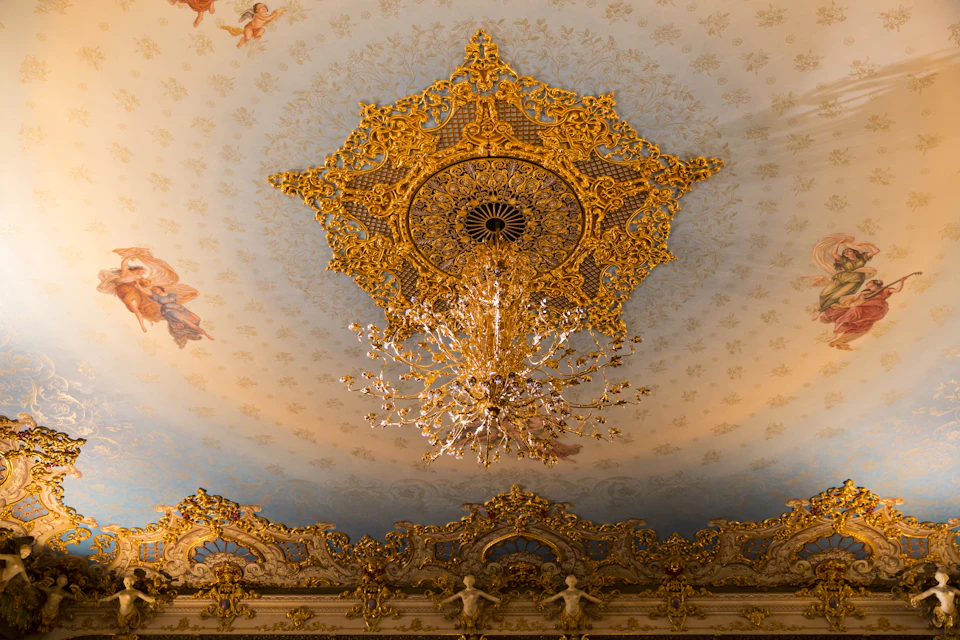 Ornate golden chandelier and intricate golden ceiling decoration in an elaborately detailed room with painted cherubs.