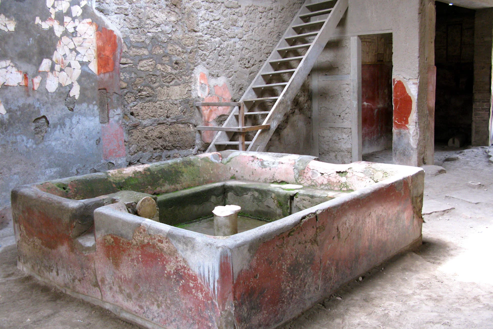 A stone basin with red and green tones in a rustic room with stairs leading up and aged plaster walls.