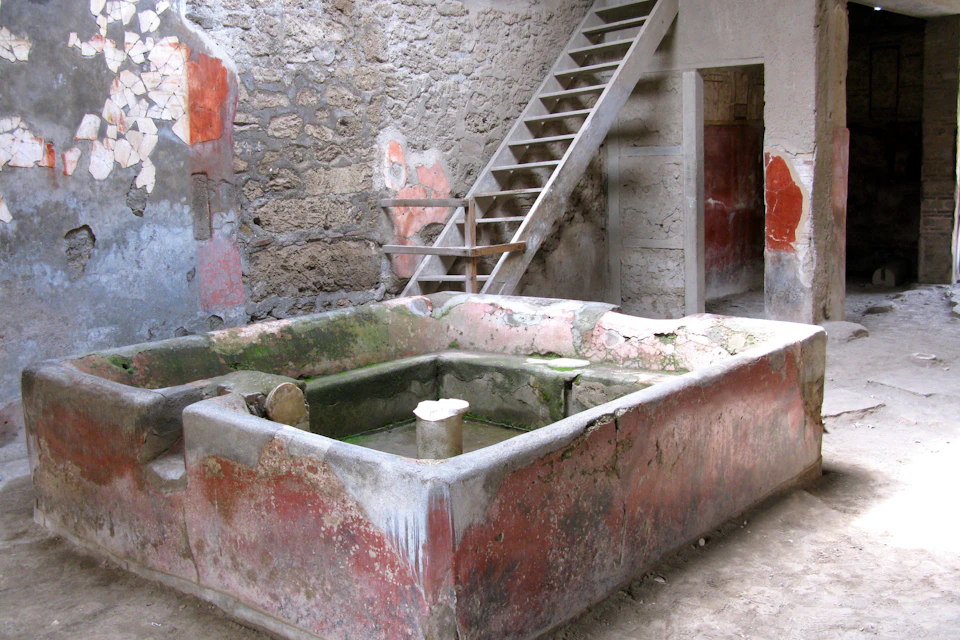 A stone basin with red and green tones in a rustic room with stairs leading up and aged plaster walls.