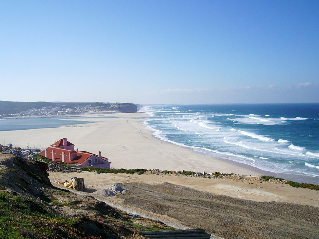 広い砂浜に波がぶつかる海岸の光景、左側に赤い屋根の家、上に澄んだ青い空。