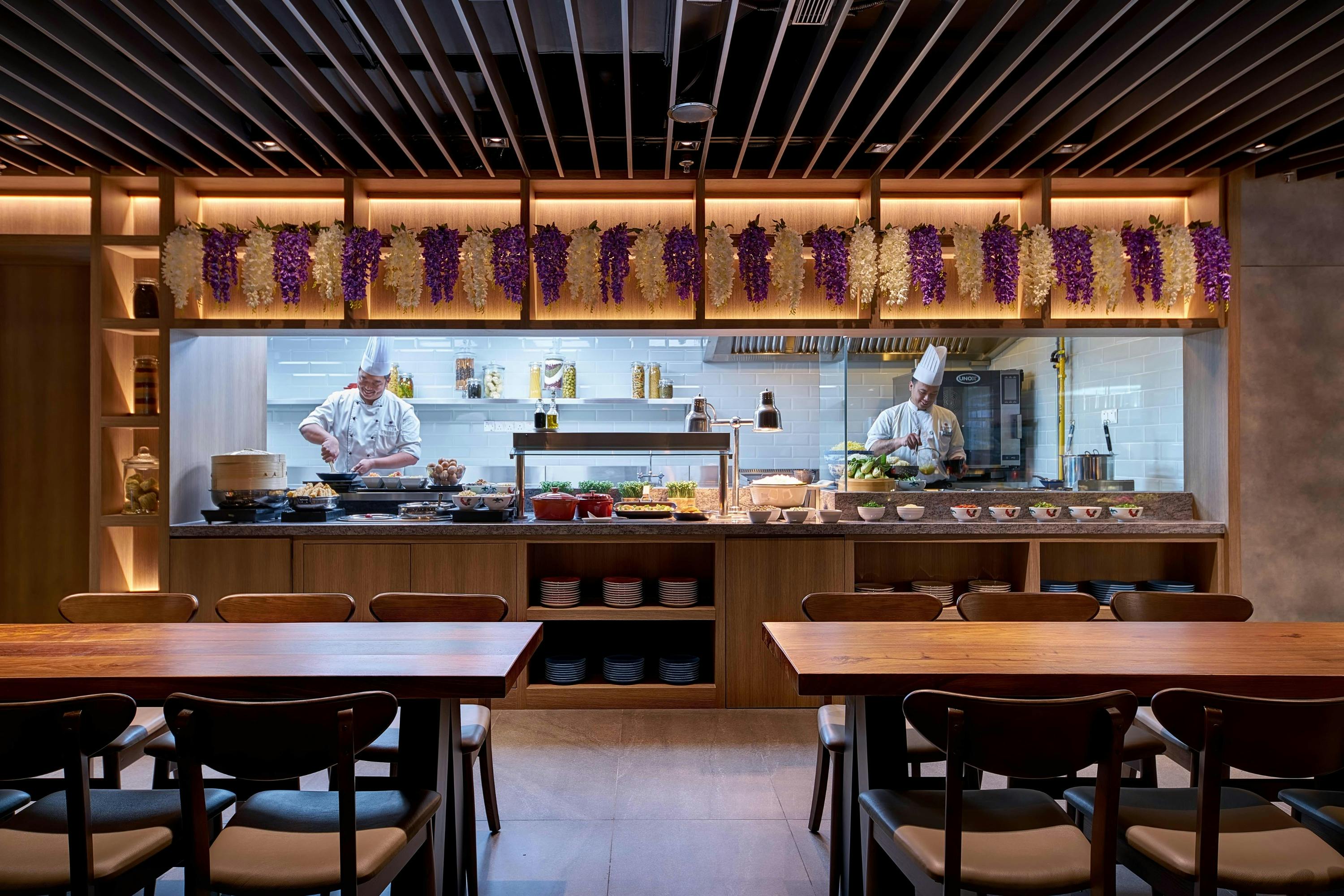 A restaurant kitchen with chefs preparing food, wooden tables and chairs in the foreground, and grape decorations above.