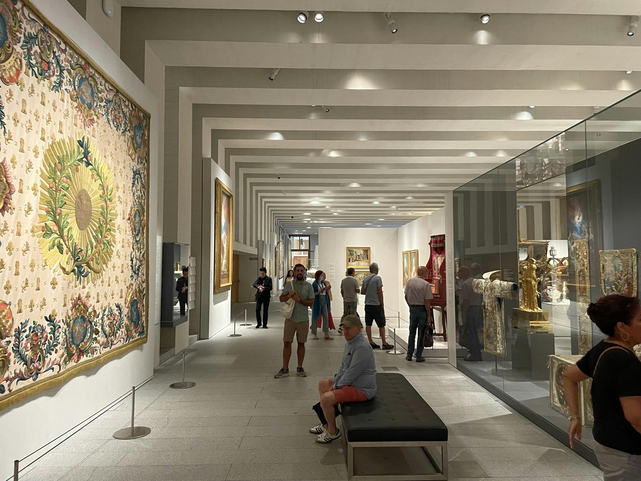 人们在光线充足的博物馆画廊里观看艺术品和文物，画廊里有挂毯、绘画和玻璃展示柜。