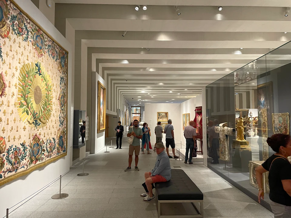 People observing artwork and artifacts in a well-lit museum gallery with a tapestry, paintings, and glass display cases.