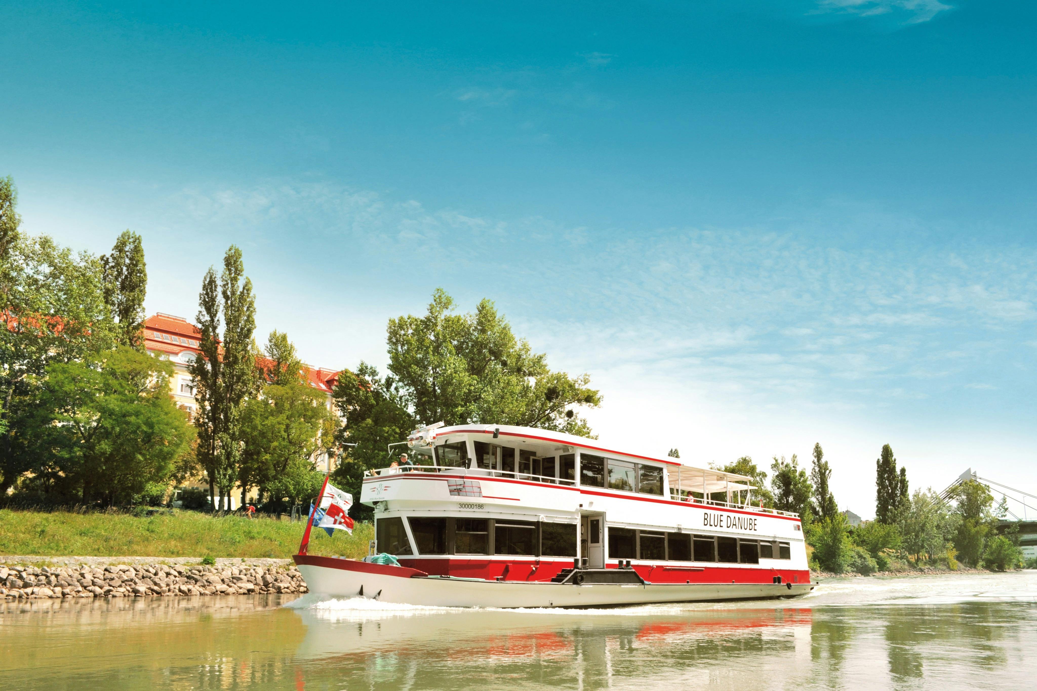 A riverboat named "Blue Danube" cruises on a river near a shoreline with trees and buildings, under a clear blue sky.