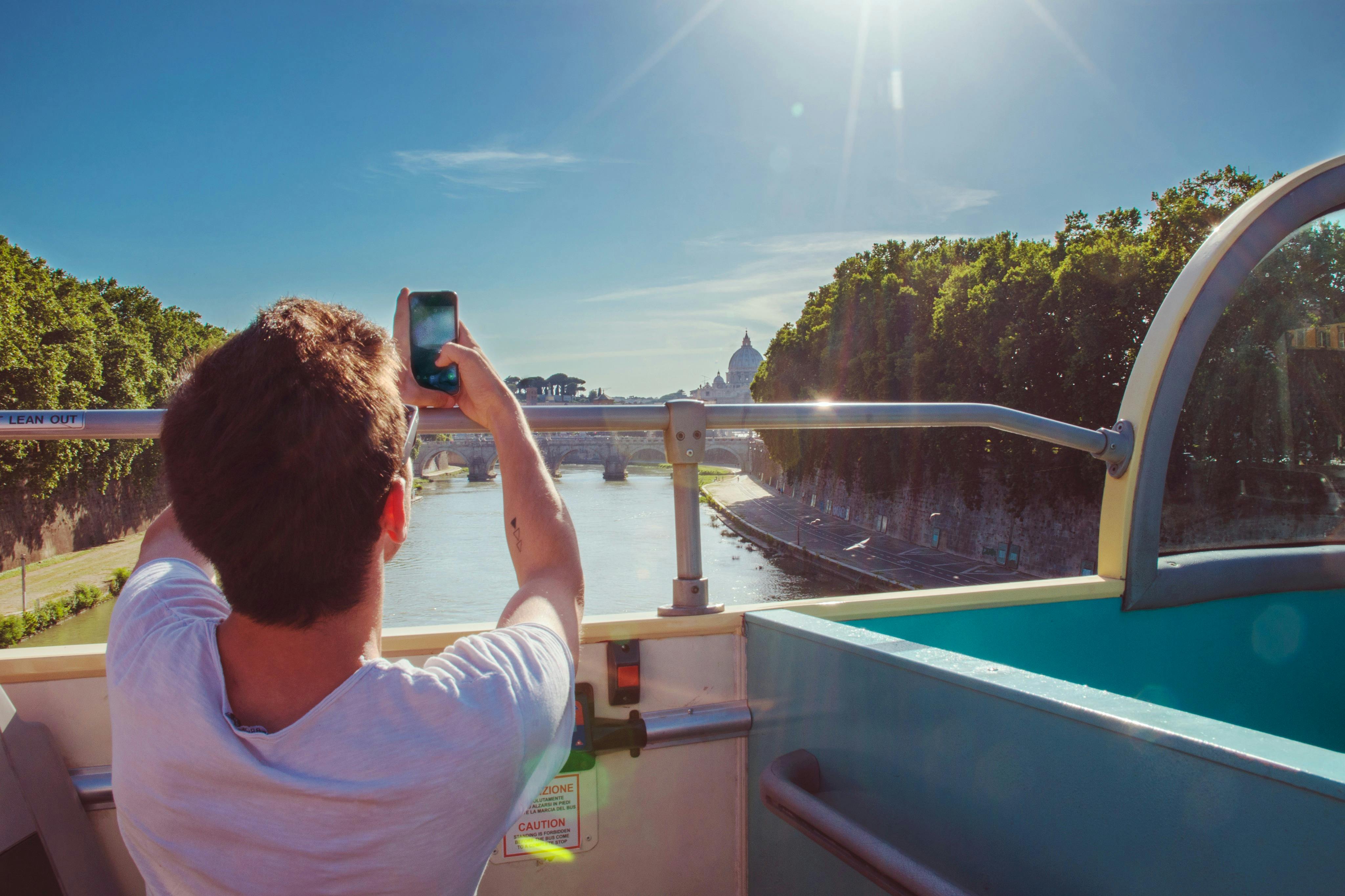Person taking a photo with a smartphone from an open-top bus, capturing a river, bridge, and distant dome under a clear sky.