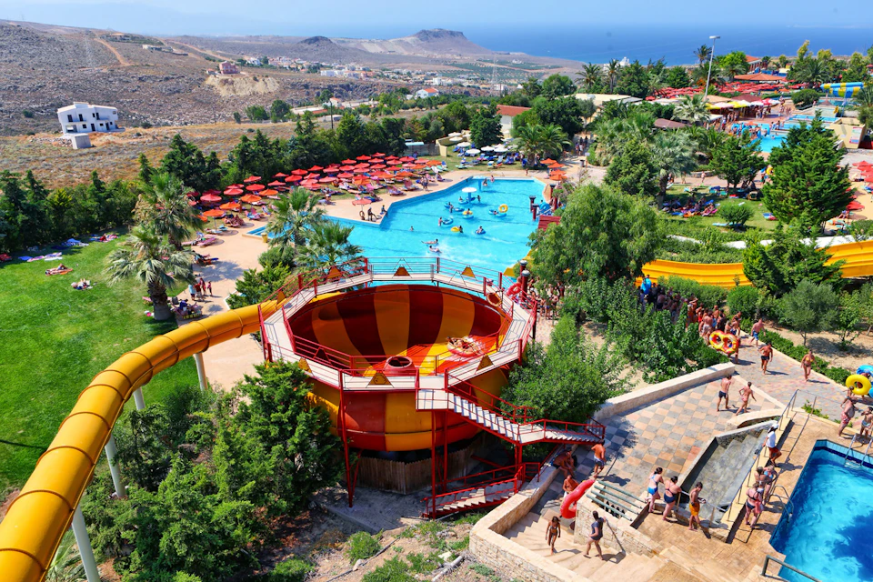 People at a water park with a large funnel water slide, pool, sunbathing area with red umbrellas, and surrounding greenery.