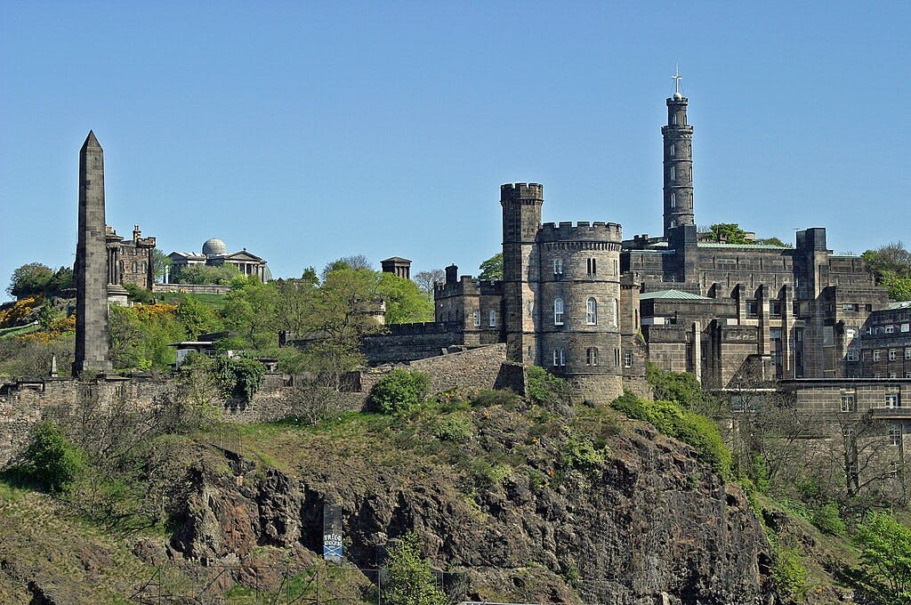 Des tours de pierre, des monuments et des bâtiments historiques se dressent sur une colline rocheuse et verdoyante, sous un ciel bleu limpide.
