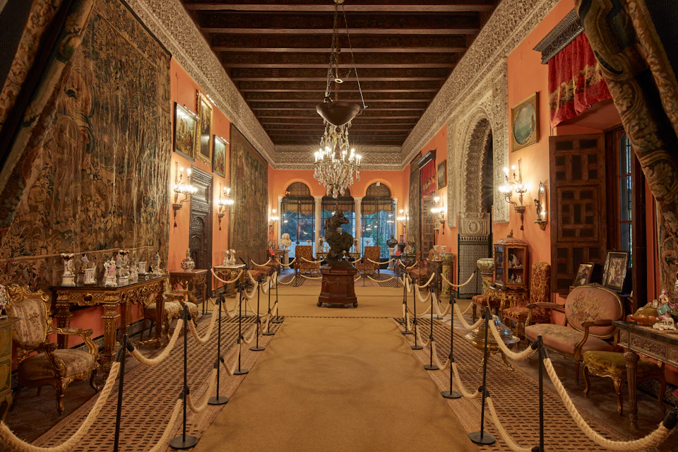 Lavish room with chandeliers, artwork, tapestries, ornate furniture, and roped-off walkway.