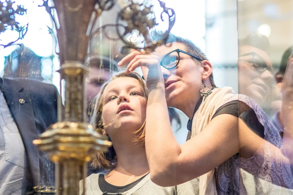 A group of people, including a child and an adult, closely observe an intricate ornate object behind glass in a museum.