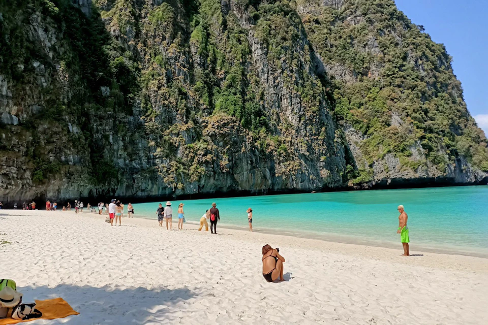 People enjoy a sunny day on a white sandy beach with turquoise waters, set against lush, rocky cliffs. One person kneels, taking a photo.