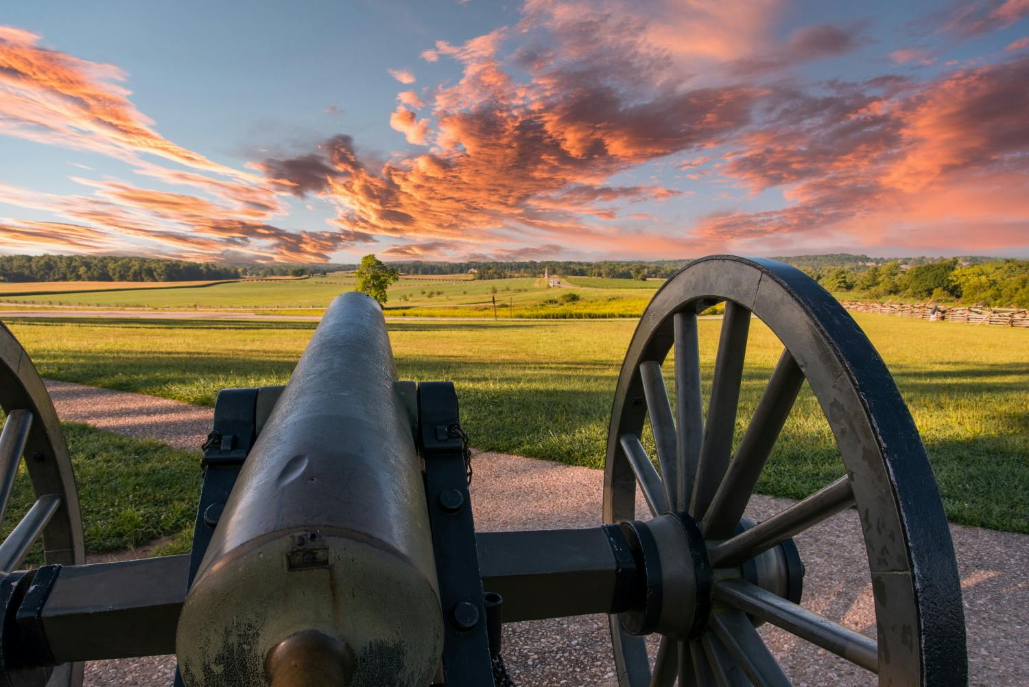 Un canon historique pointe vers des champs ouverts sous un ciel dramatique avec des nuages roses et vibrants au coucher du soleil.