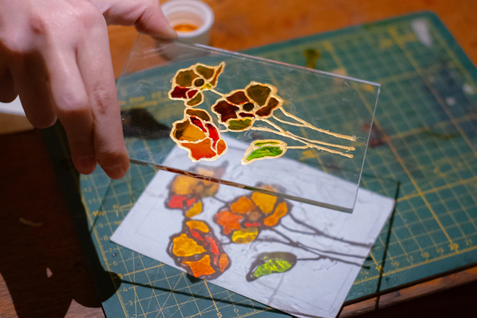 A person holds a glass sheet with colorful painted flowers, above a matching design sketch on a cutting mat.
