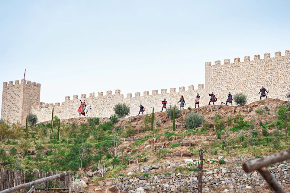 A group of people dressed in medieval-style clothing perform on a hillside with a stone wall in the background.