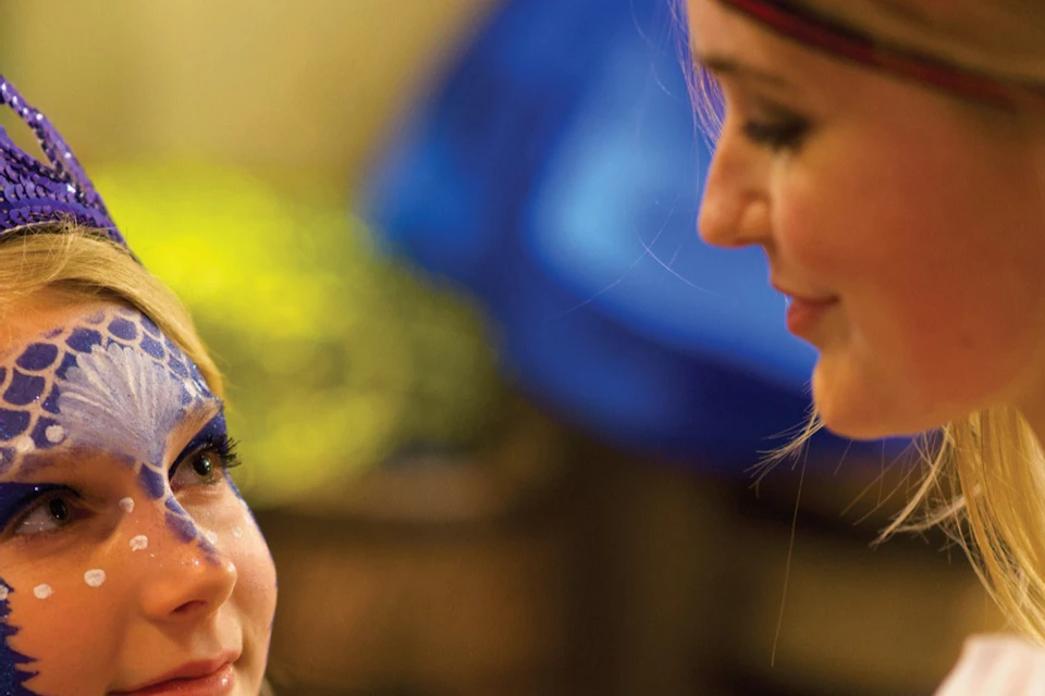 A woman paints a colorful design on a girl's face, who is wearing a blue tiara, in a warmly lit indoor setting.