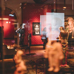 Ett museum ställer ut med besökare, porträttmålningar på röda väggar och visar skulpturer genom en glasreflektion.