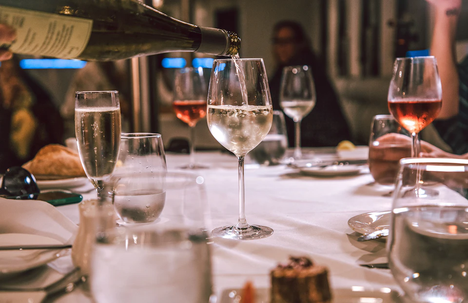 Wine being poured into a glass on a dining table set with glasses, plates, and a dessert in the foreground.