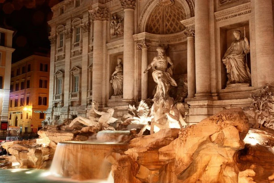 Illuminated marble statues and cascading water of the Trevi Fountain at night, with ornate architectural details in the background.