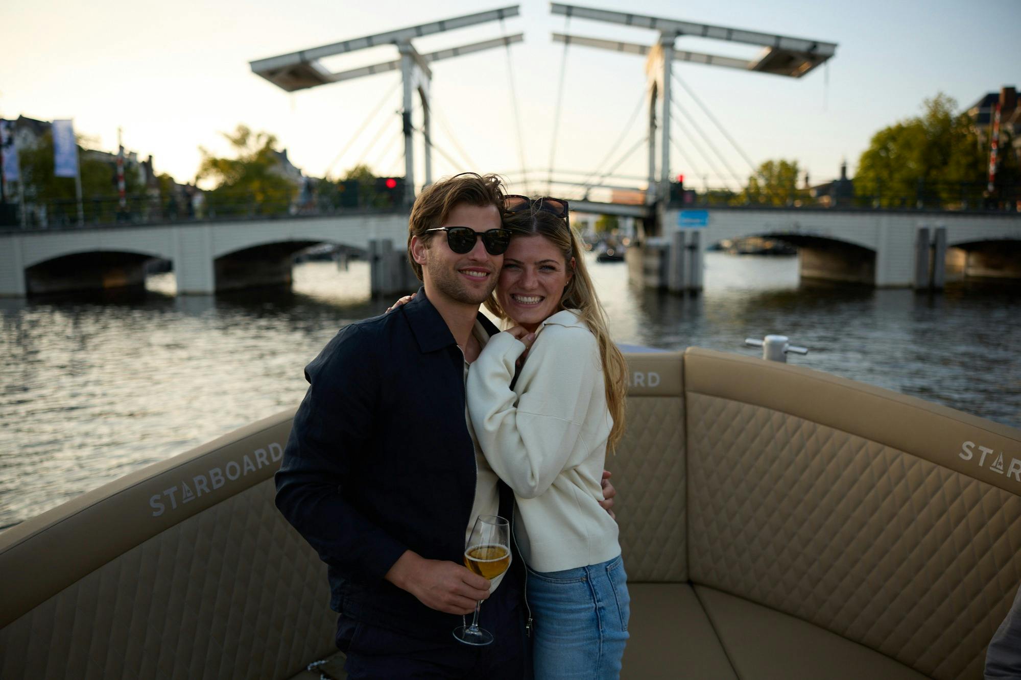 グラス1杯のワインを片手に、運河と橋が見えるボートの上で、笑顔のカップルがポーズを取っています。