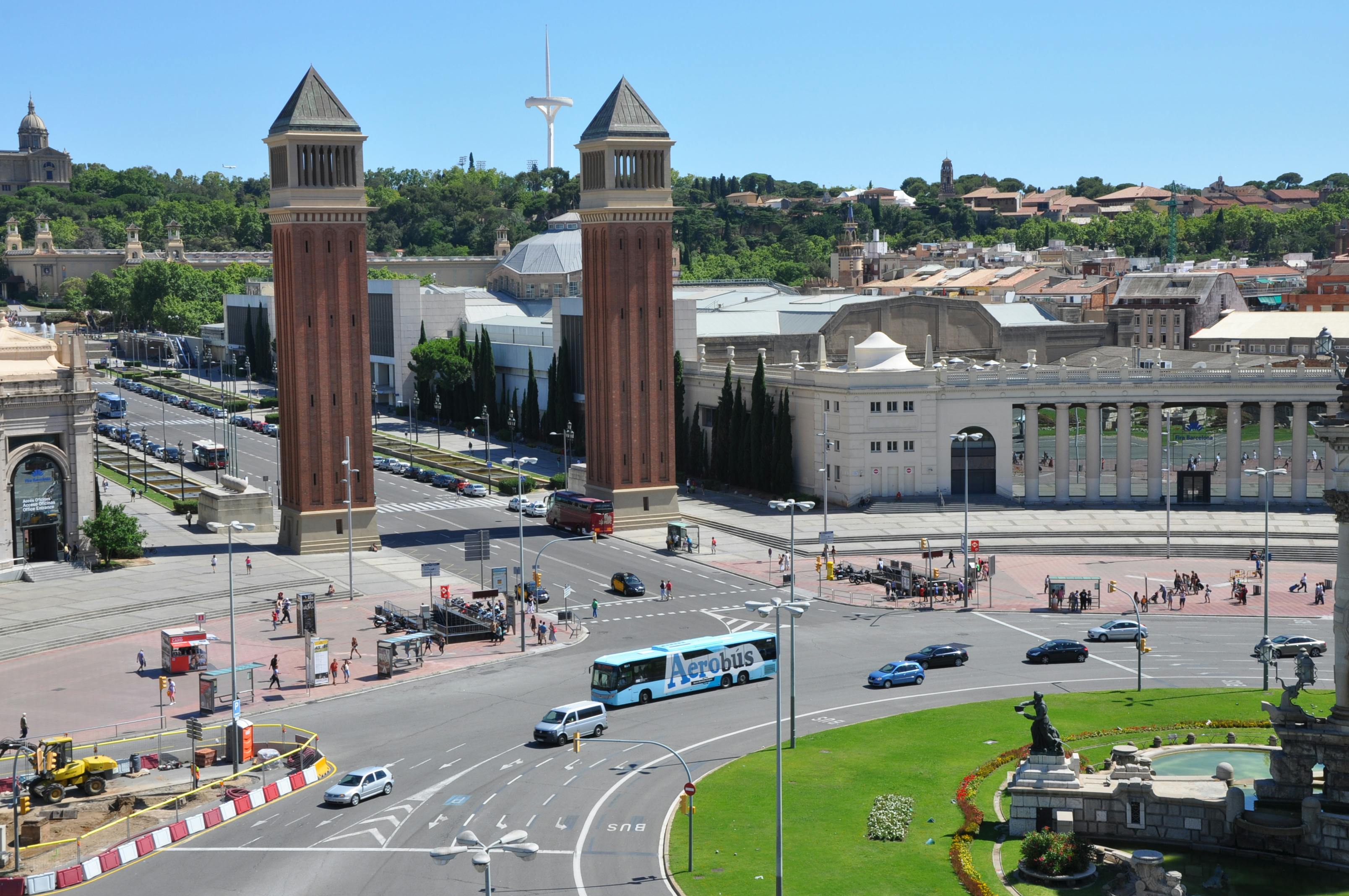 Scena urbana con due alte torri di mattoni, una rotonda trafficata con auto, un Aerobus blu ed edifici storici circostanti.